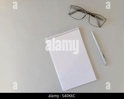 Flat display of a notepad and pen with a pair of glasses on a plain light grey background. Copy space. Stock Photo