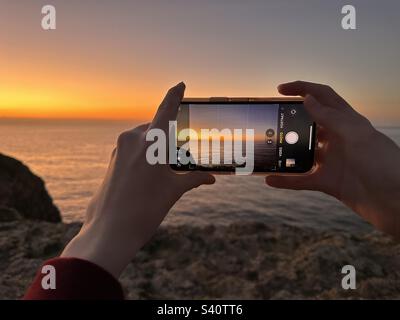 Hands holding mobile phone while taking photo of sunset Stock Photo