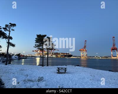 Halifax port in winter sunset: snow-covered docked ship with twinkling lights, serene orange-pink sky reflects on calm waters, distant boats, and city skyline create a peaceful and resilient scene Stock Photo