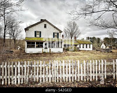 White farmhouse in Lebanon, Connecticut, that is falling apart, and actually has moss growing on the roof. White picket fence in front. Stock Photo