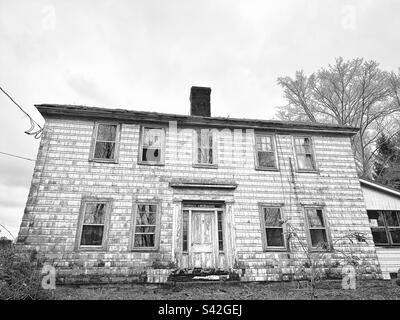 Black-and-white photo of an old, rundown white farmhouse in Lebanon, Connecticut during winter. The exterior looks dirty, and is deteriorating. Stock Photo