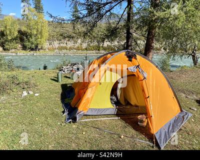 An orange extreme tourist tent with equipment and drying socks stands on the bank of a mountain river in Altai. Stock Photo