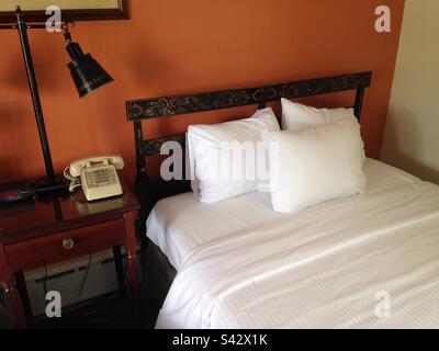 Hotel bedroom in Vermont with white sheets and old fashion, ornate black headboard with beautiful, floral detail. Older style telephone with push buttons. Also, orange accent wall. Stock Photo