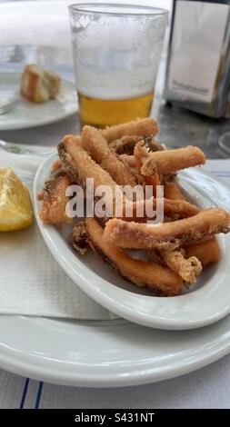 A small plate of fried “rabas” or calamari strips, a lemon wedge, a half empty grass of beer a paper napkin holder, a chuck of baguette bread, tapas appetizers in north Spain Stock Photo