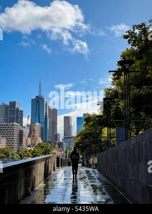 Rear view of man walking along paved footpath against buildings and city skyline after the rain in Melbourne, Australia. Stock Photo