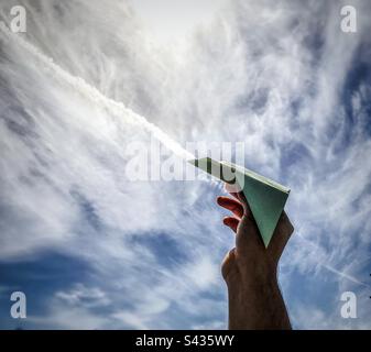 Human hand holding paper airplane against vapor trail in dramatic, cloudy, blue sky. Stock Photo