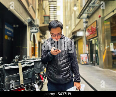 Young Asian man using smartphone while standing beside food delivery bicycles in laneway against buildings in the city in Melbourne, Australia. Stock Photo