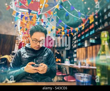 Young Asian man using mobile phone at table inside restaurant decorated with colorful buntings. Stock Photo