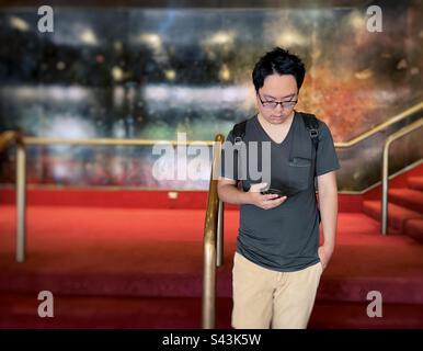 Young Asian man using smartphone on staircase against railings and wall. Stock Photo
