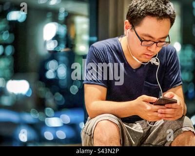 Young Asian man with earphones and eyeglasses using mobile phone against defocused reflections in window at night. Stock Photo
