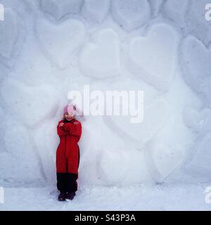 A young 6-year-old girl in front of a snow heart wall in a public play park in deep winter in Scandinavia Stock Photo