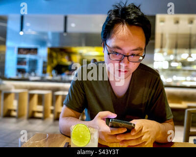 Young Asian man in eyeglasses using smartphone at table inside restaurant. Wireless technology. Communication. Stock Photo