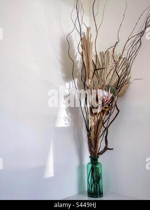 Arrangement of dried flower, palm leaf, grasses and branches in green glass bottle on white table against white wall with shadow patterns. Art. Stock Photo