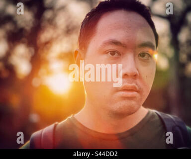 In the gloaming. Close-up headshot of young Asian man against trees and setting sun with lens flare. Focus on foreground. Back lit. Stock Photo