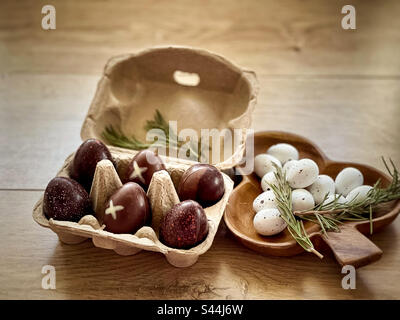 High angle view of chocolate Easter eggs in cardboard egg carton and praline Easter eggs in decorative wooden container with sprigs of rosemary on wooden table. Rustic style. Easter. Celebration. Stock Photo