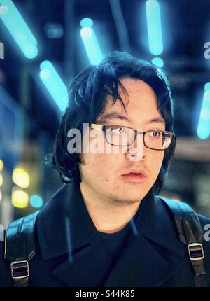 Close-up portrait of young Asian man in eyeglasses standing against blue neon lights in the city at night. Stock Photo