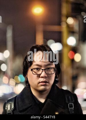 Portrait of young Asian man in eyeglasses against street lights and building in the city at night. Focus on foreground. Stock Photo