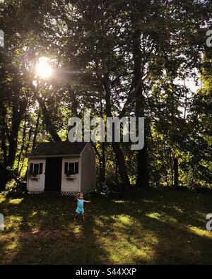 Little girl runs across green Cape Cod lawn with a little cottage in the background as light shines through trees. Stock Photo