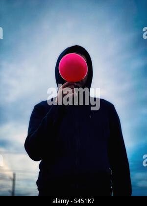 Surreal man in black hood with red balloon obscuring his face wandering against glowing cloudy blue sky. Dreamscape. Archetype. Wanderer. Explorer. Abstract. Stock Photo
