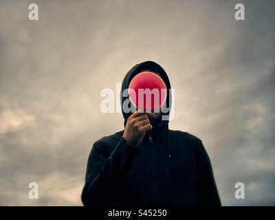 Surreal man in black hoodie holding red balloon obscuring his face, standing against glowing cloudy sky. Dreamscape. Dystopian. Stock Photo