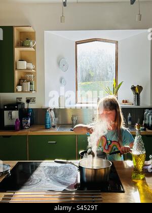 Girl cooking on a convection hob, stirring a pan with steam coming out. Window behind in an open plan kitchen Stock Photo