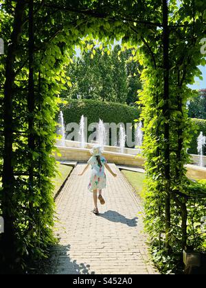 Girl running towards fountains in the Alnwick garden, seen through a framed archway window of leaves Stock Photo