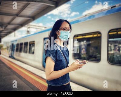 Young Asian woman in eyeglasses and face mask using mobile phone at railway station. Stock Photo