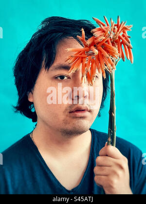 Close-up portrait of young Asian man holding a stalk of orange aloe vera flower against blue background. Stock Photo