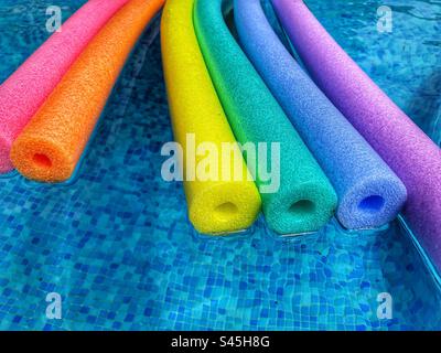 Summer vibes, rainbow coloured polyethylene pool noodles floating on water of blue tiled swimming pool, with copy space Stock Photo