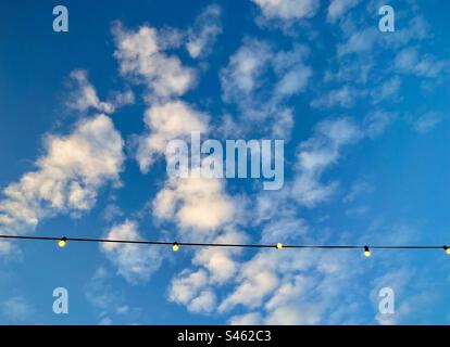 Low angle view of glowing light bulbs hanging from overhead line against blue sky with fluffy white clouds with copy space. String of light bulbs. Stock Photo