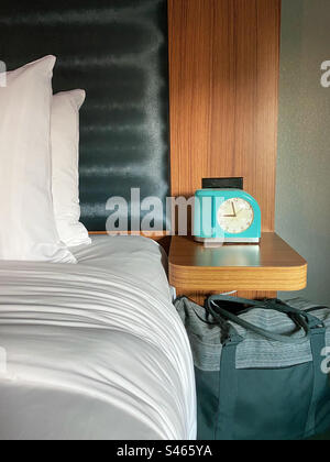 Hotel room bed and bedside table with vintage alarm clock and overnight bag. Stock Photo