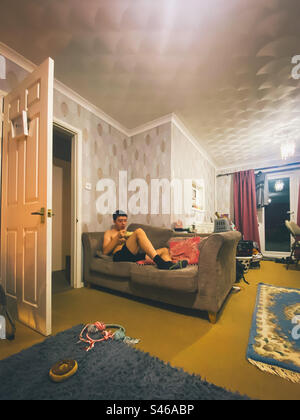 A teenage boy sat on a sofa in the evening eating a bowl of cereal. Stock Photo