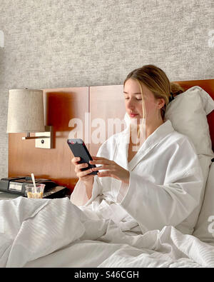 Young beautiful blonde woman dressed in bathrobe while sitting in hotel room bed using mobile phone. Stock Photo