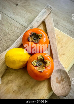 High angle view of a lemon and two persimmons together with two wooden spoons on wooden cutting board on table. Rustic look. Healthy eating. Stock Photo
