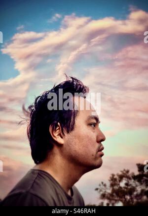 Portrait of young Asian man lost in contemplation against pink sunset clouds in blue sky. Wind. Windswept clouds. Stock Photo