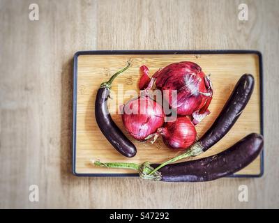 Directly above view of three red onions and three Lebanese eggplants on wooden cutting board on wooden table with copy space. Stock Photo