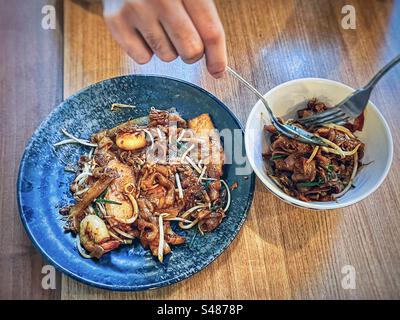 Man’s hand using fork and spoon to scoop up char koay teow/ spicy, fried flat noodles with prawns, mung bean sprouts and chives into a bowl on table. Malaysian food. Stock Photo