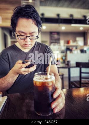 Asian man in eyeglasses using a mobile phone while drinking iced coffee at table in a cafe. Wireless technology. Communication. Food and drink. Stock Photo