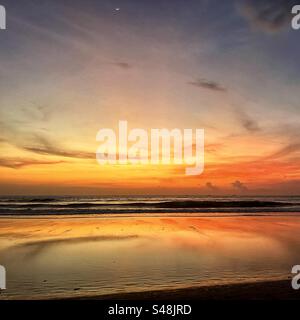 Sunset at Seminyak beach Bali with the sun set over the sea and the orange glow remaining in the clouds and the orange light reflecting in the shallow water Stock Photo