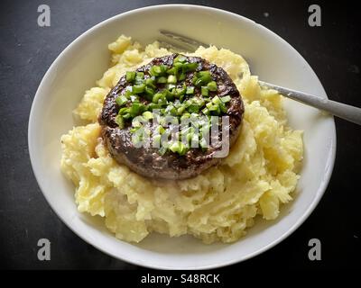 Shot of a white round plate of hamburger steak with green onions sauce on a bed of homemade mashed potatoes with butter and a fork on background of black wooden table Stock Photo