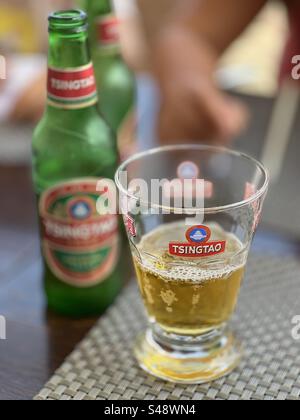 A glass of Tsing-Tao beer next to its empty bottle on a bar restaurant table in Palavas, Occitanie, France Stock Photo
