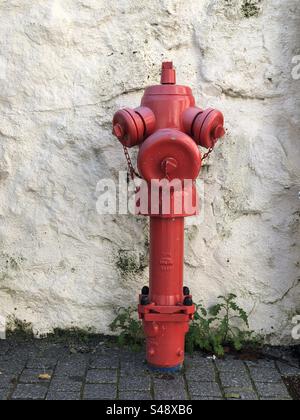 Viana do Castelo, Portugal - December 01, 2023: A vibrant red fire hydrant with chains stands against a textured white wall on a cobblestone street Stock Photo