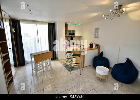 Interior shot of a bright Airbnb studio apartment with table and chairs, open concept American kitchen and white floor tiles. Stock Photo