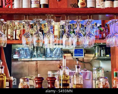 Empty wine glasses hanging down in front of bottles of hard liquor on shelves at the counter of an Italian restaurant with condiments and olive oil bottles. Stock Photo