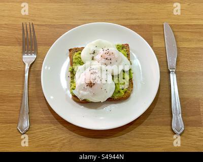 Poached eggs on avocado toast sprinkled with black pepper on a white plate between a knife and fork. Stock Photo