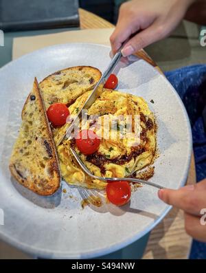 Hands holding knife and fork over chili scrambled eggs with cherry tomatoes and sourdough toast on plate on table. Healthy breakfast. Stock Photo