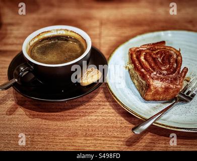 Cinnamon scroll on plate with fork and a cup of black coffee on saucer with small biscuit on wooden table. Coffee break. Stock Photo