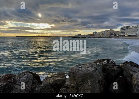 Landscape shot of the sun setting through the clouds over the waves, with a rocky pier on the foreground, on the Mediterranean in Palavas-les-Flots, Occitanie, France. Stock Photo