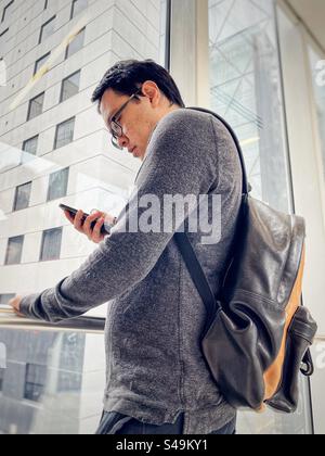 Low angle view of Asian man with backpack using mobile phone against window view of buildings in the city. Stock Photo