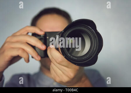Young man using a professional camera. Close up photo of man in hat on blue background taking a photo with digital mirrorless camera. Paparazzi taking Stock Photo